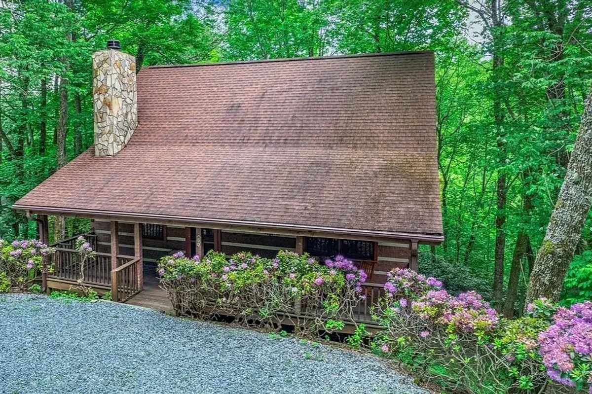 An inviting exterior view of the cabin's stone chimney and wooden porch, surrounded by lush greenery and vibrant flowering bushes. The porch area hints at opportunities for morning coffee, evening relaxation, and soaking in the fresh mountain air, all within steps of the region's adventure attractions.