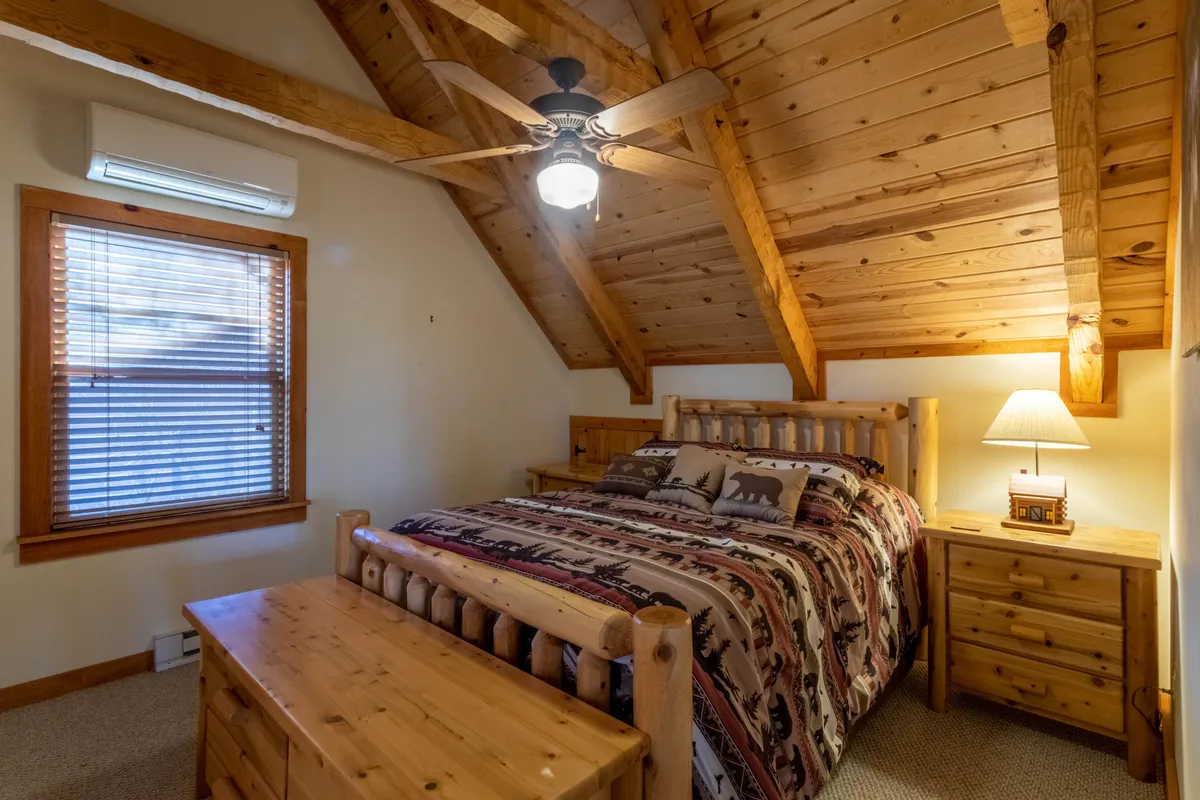 This image features a wooden bed frame adorned with decorative pillows, a ceiling fan, and a nightstand with a lamp, all set in a warmly lit, inviting space. The styling suggests a serene environment, ideal for couples or grandparents seeking privacy and comfort in the upstairs queen suite.