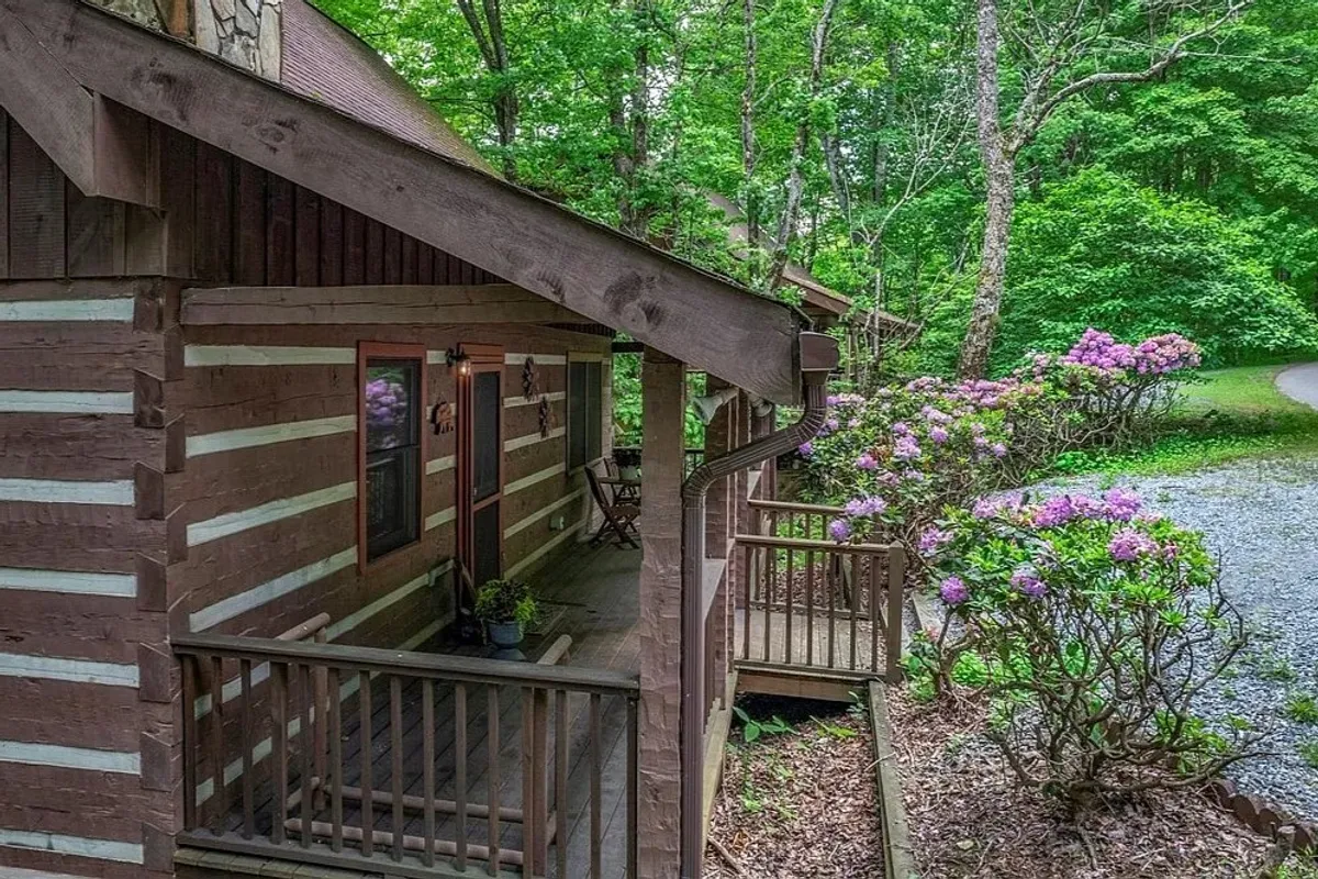 This image showcases the exterior of the cabin, with a classic log cabin structure nestled among flowering bushes and a wooden deck. The inviting porch is visible, surrounded by natural greenery, evoking a sense of tranquility and mountain charm.
