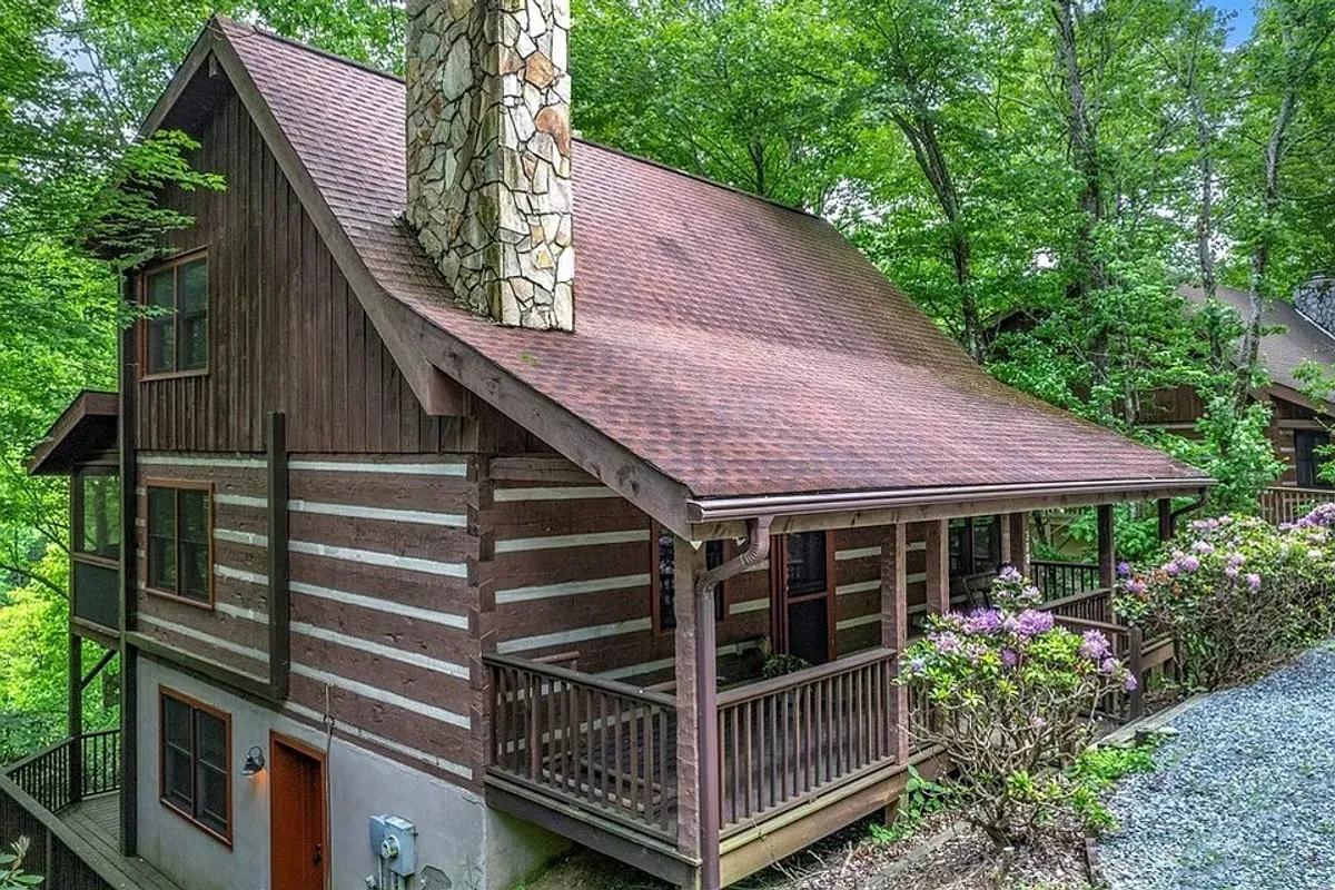 The image showcases a classic log cabin with a rustic stone chimney, inviting front porch, and is surrounded by vibrant green forest. The tranquil setting suggests a peaceful escape, with sunset views and ample space for guests to relax and enjoy nature.