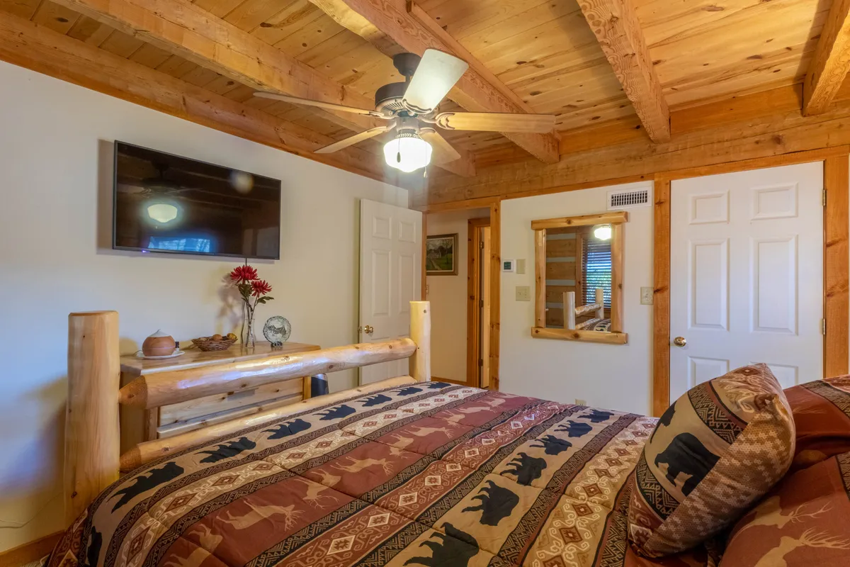 A cozy bedroom with a sturdy log bed frame, decorative pillows, ceiling fan, television, and rich wooden accents. The space feels restful and inviting, providing a tranquil retreat adjacent to the loft.