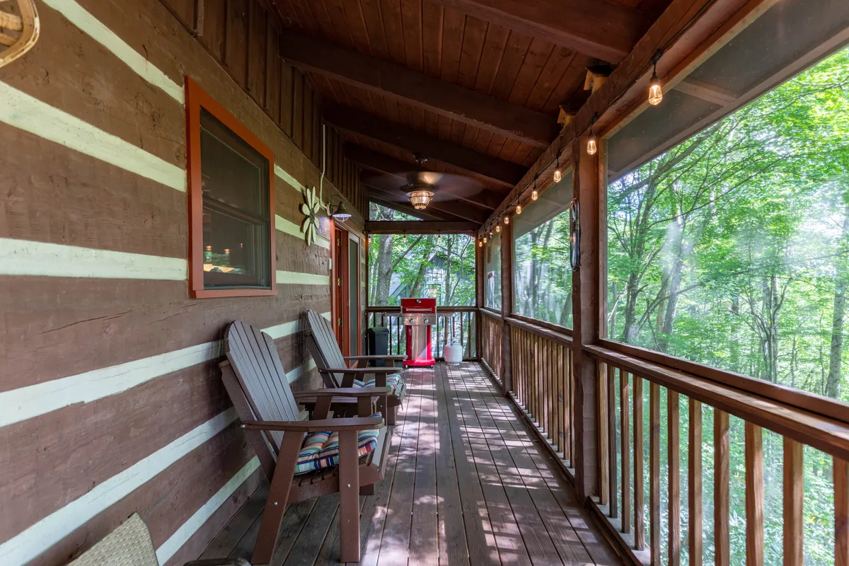 This image captures a spacious wooden deck surrounded by trees, featuring rocking chairs, a grill, and string lights. The inviting outdoor space is designed for relaxation, socializing, and family barbecues, with nature providing a tranquil backdrop.