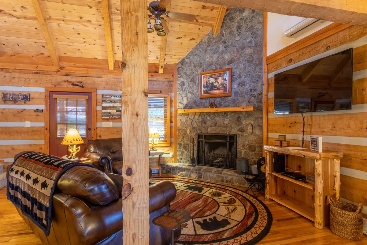 A cozy living room featuring a grand stone fireplace flanked by wooden beams, plush leather sofa, a decorative rug, and a television. The room exudes warmth and invites gatherings, with a crackling fire and views that hint at the mountain surroundings.