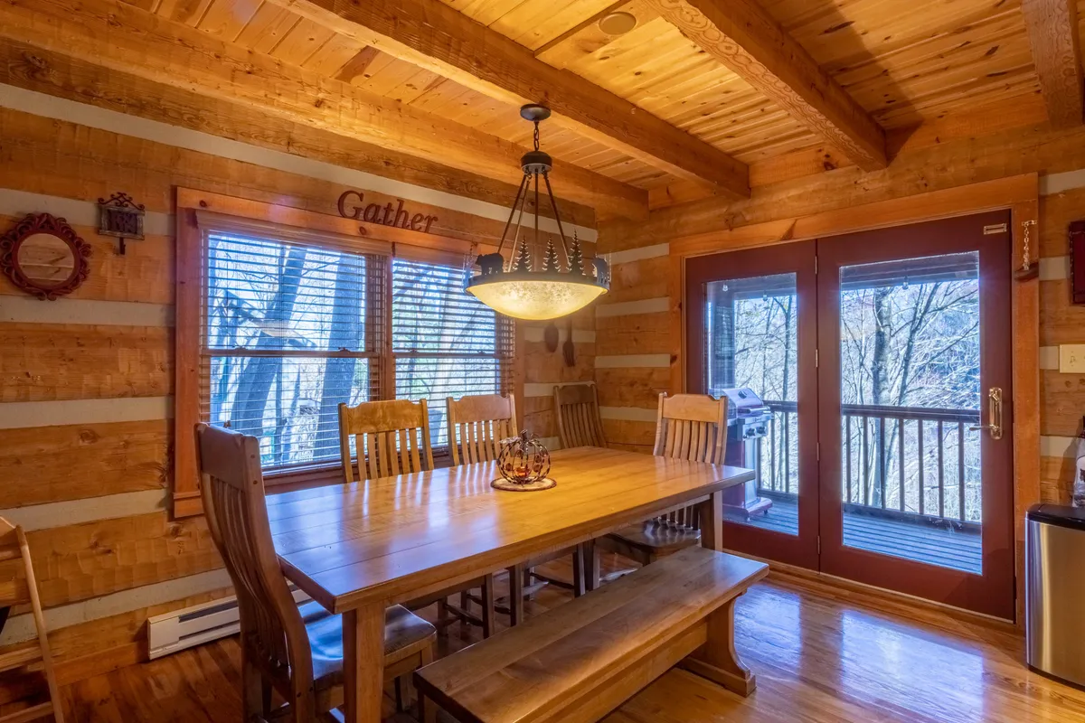 A spacious dining area centered around a large wooden farmhouse table under a chandelier, surrounded by wooden beams and inviting natural light from sliding glass doors. The ambiance is welcoming and perfect for group meals or games.